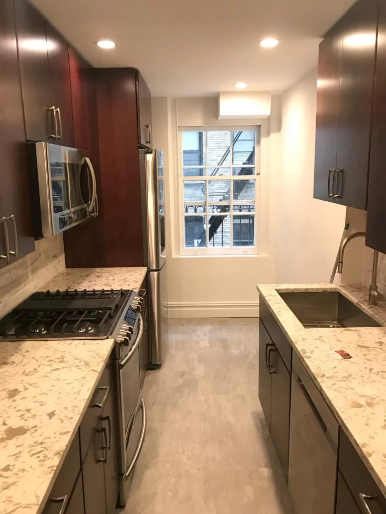 A narrow kitchen with dark wood cabinets, granite countertops, a gas stove, and a window showing scaffolding outside, possibly for home remodeling services.
