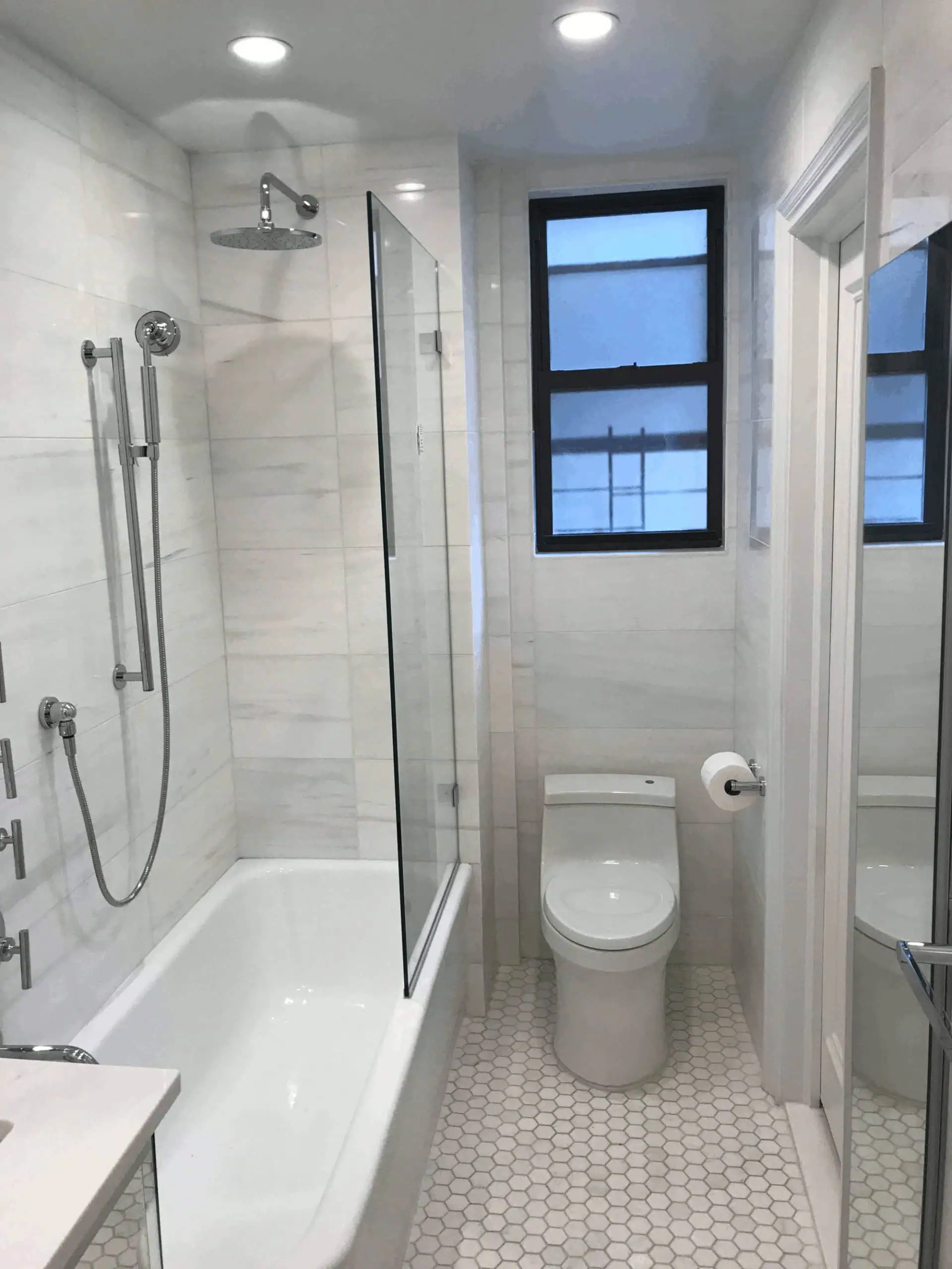 Modern bathroom renovated by MHI Renovation Services Inc, featuring a white bathtub, glass shower partition, toilet, and a window with a dark frame.