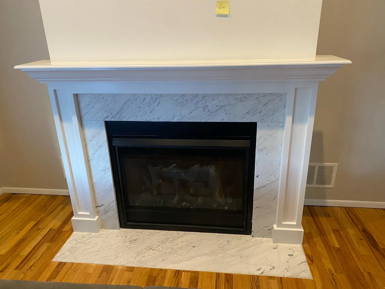 A traditional white fireplace with a black insert and faux logs, set against a marble backdrop and hardwood flooring, suitable for home remodeling projects.