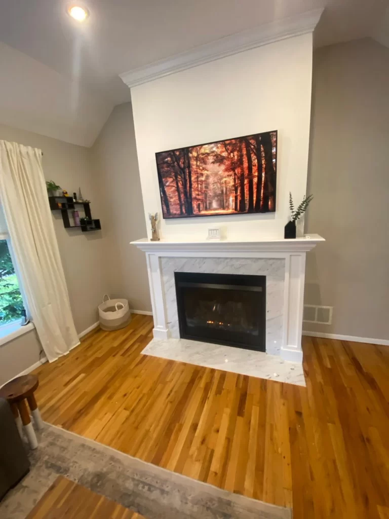 Cozy living room with a white fireplace under a wall-mounted painting of a forest, flanked by a window with white curtains and wood flooring, enhanced by home remodeling services.