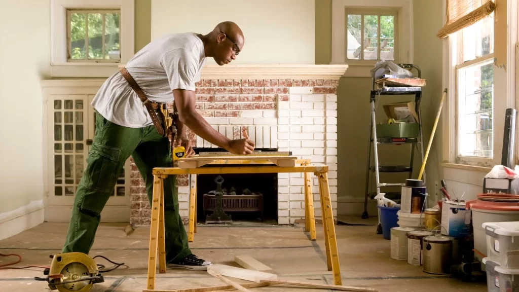 A man in a white shirt and green pants measuring wood on a sawhorse in a room undergoing renovation services.