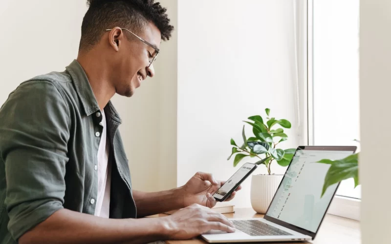 A smiling man uses a smartphone and laptop at a desk near a window to get a quote, with a plant beside him.