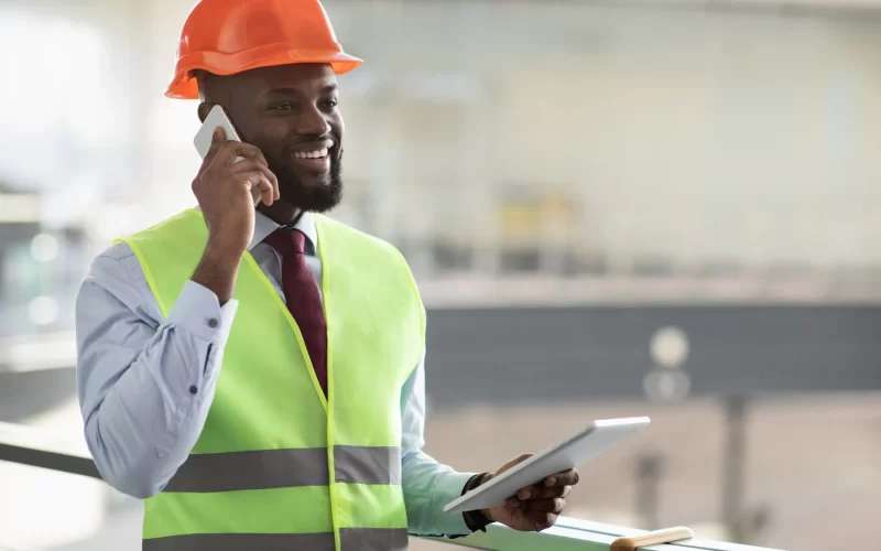 A construction manager specializing in home remodeling services, wearing a hard hat and high visibility vest, talks on a mobile phone while holding a tablet.