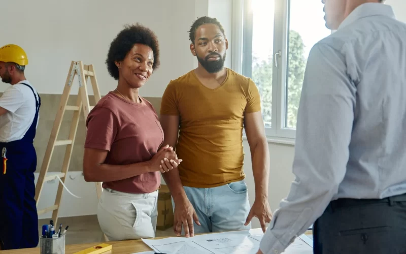 A couple reviews plans with an architect in a bright workspace, with a worker measuring in the background.